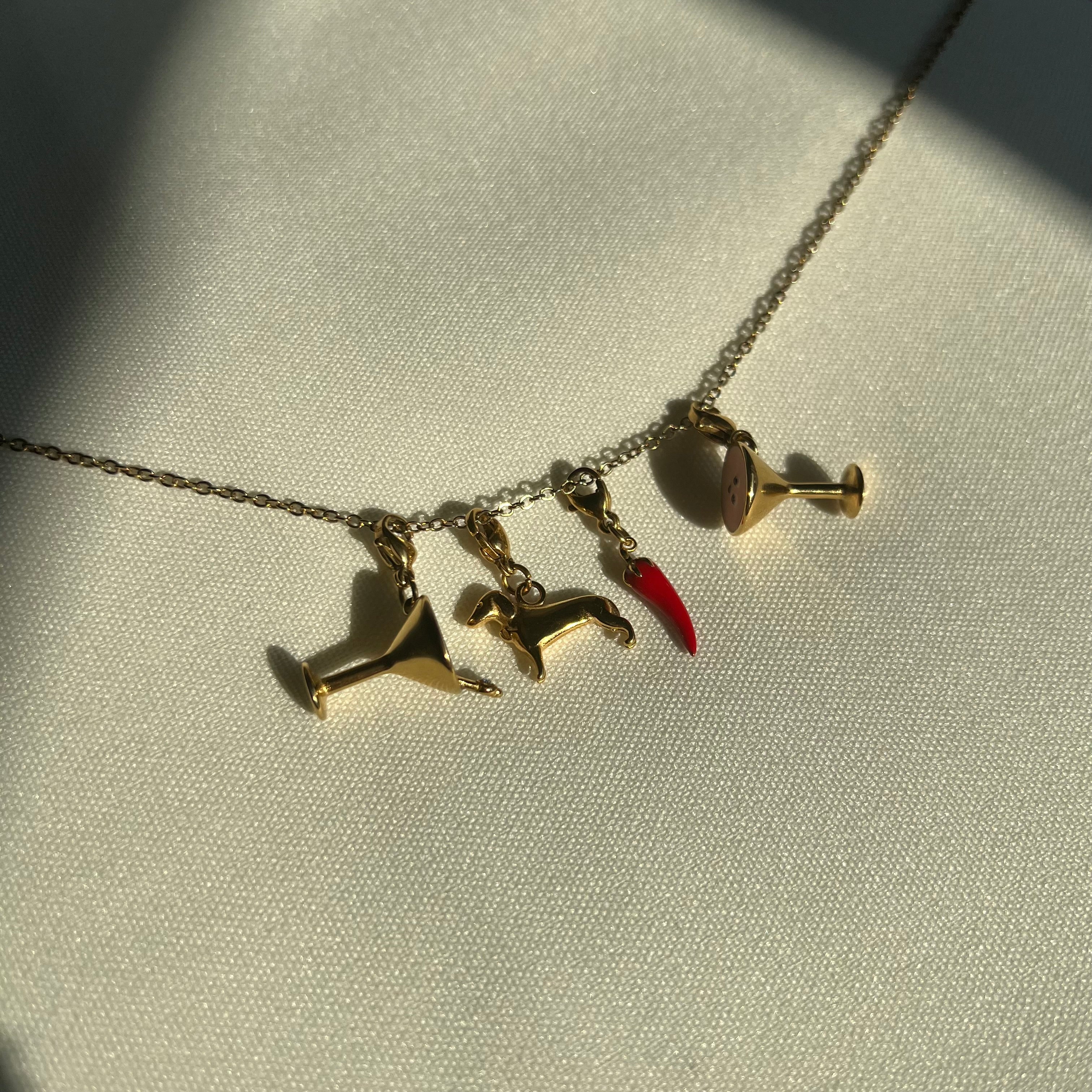 Goldener Cocktail Charm, Hunde Charm und rot goldener Chili Charm an Goldener Kette auf weißem Leinentuch. 