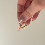 Ein goldener Espresso Martini Charm wird in den Fingern gehalten und baumelt vor weißem Hintergrund. 