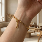 Charm B in gold hängt an goldenem Armband am Handgelenk. Im Hintergrund stylische Altbauwohnung