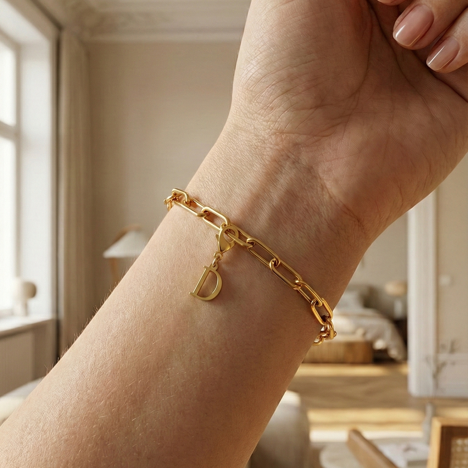 Charm D in gold hängt an goldenem Armband am Handgelenk. Im Hintergrund stylische Altbauwohnung