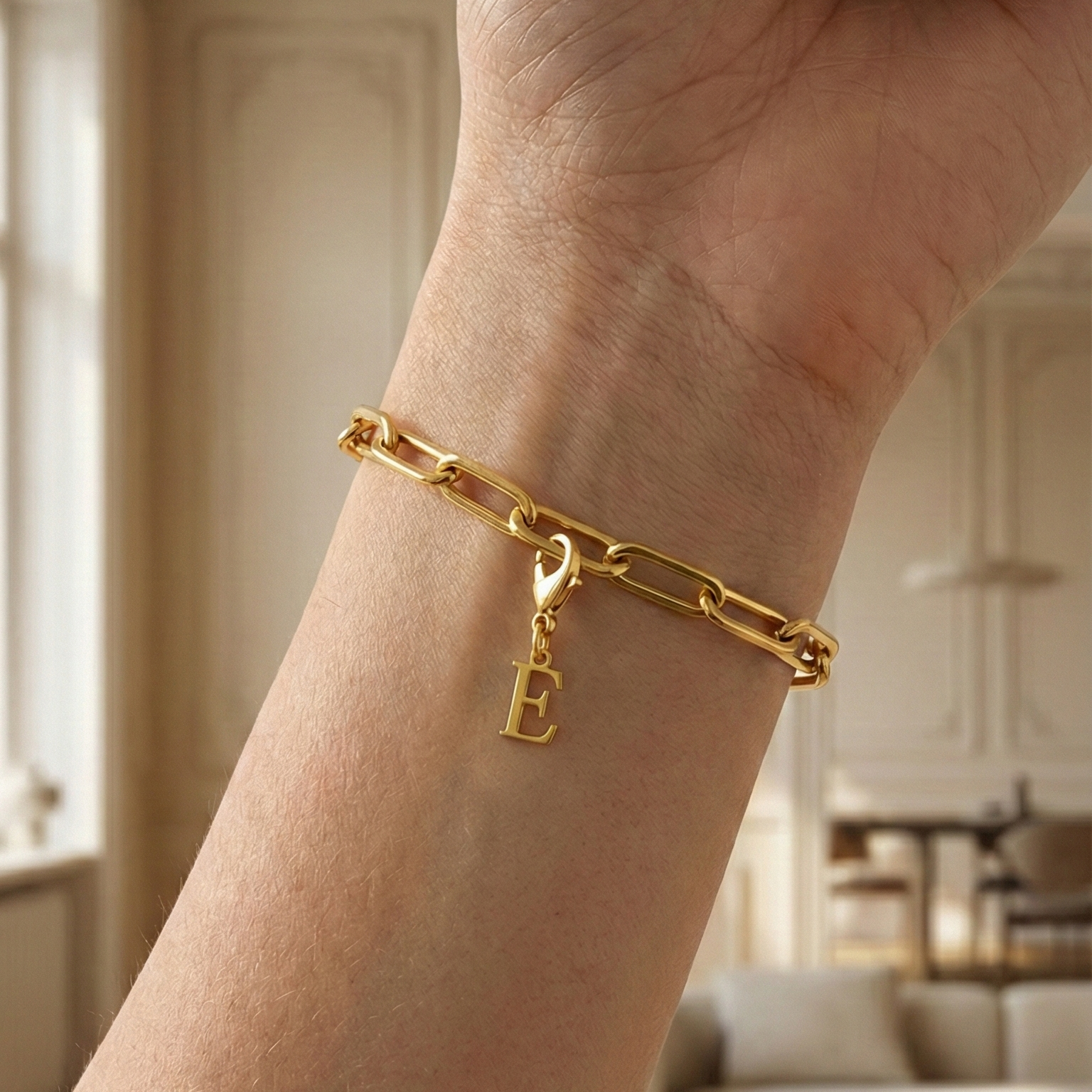 Charm E in gold hängt an goldenem Armband am Handgelenk. Im Hintergrund stylische Altbauwohnung