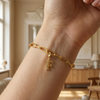 Charm F in gold hängt an goldenem Armband am Handgelenk. Im Hintergrund stylische Altbauwohnung