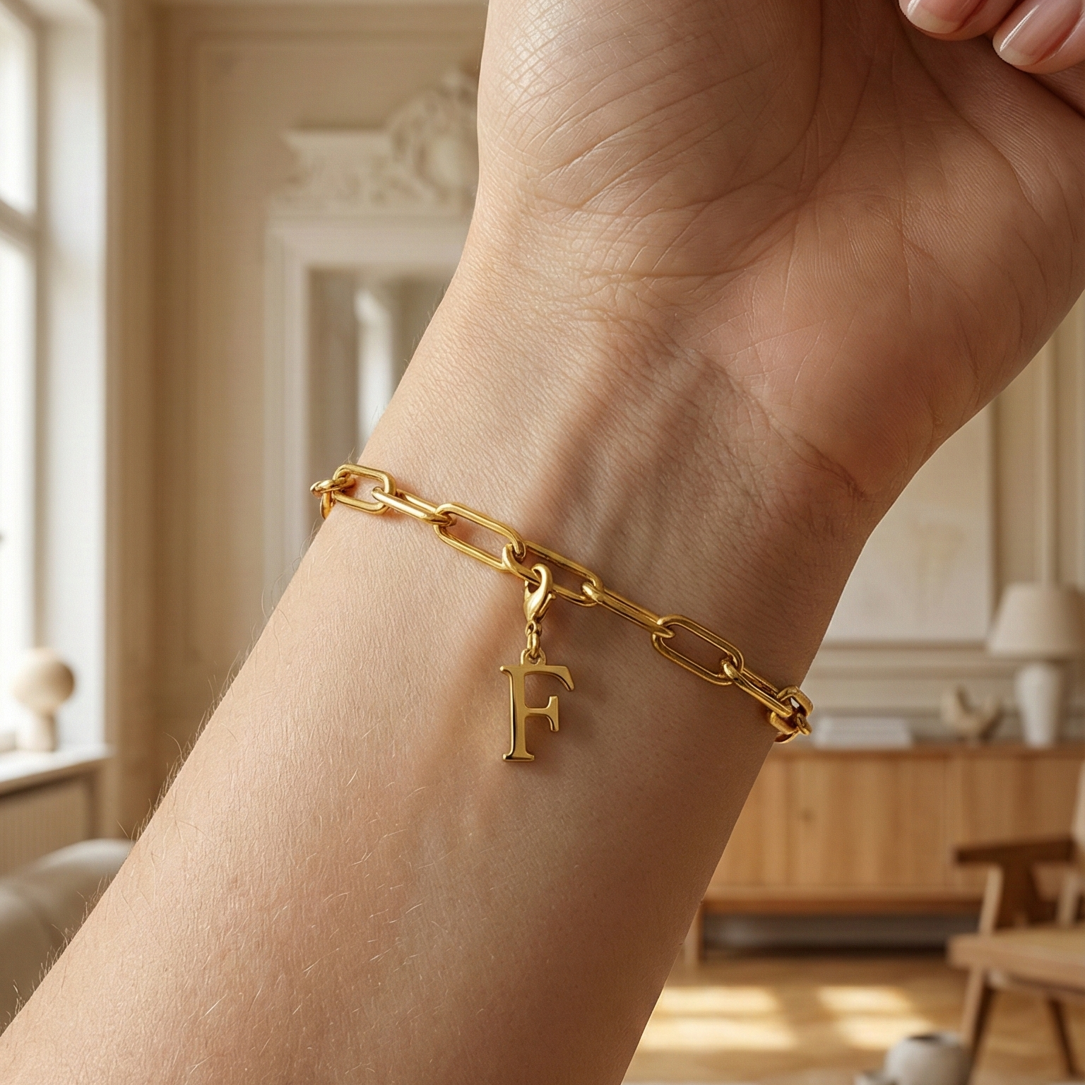 Charm F in gold hängt an goldenem Armband am Handgelenk. Im Hintergrund stylische Altbauwohnung