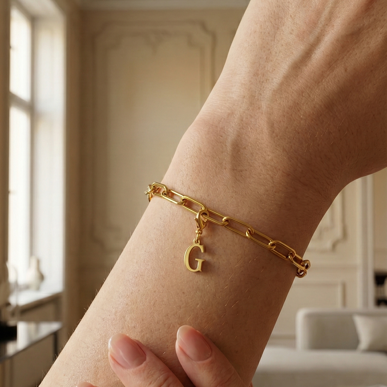 Charm G in gold hängt an goldenem Armband am Handgelenk. Im Hintergrund stylische Altbauwohnung