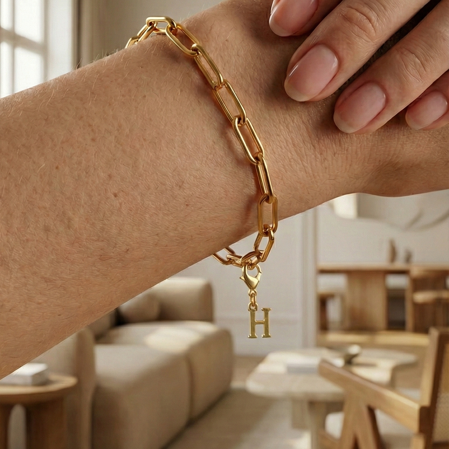 Charm H in gold hängt an goldenem Armband am Handgelenk. Im Hintergrund stylische Altbauwohnung