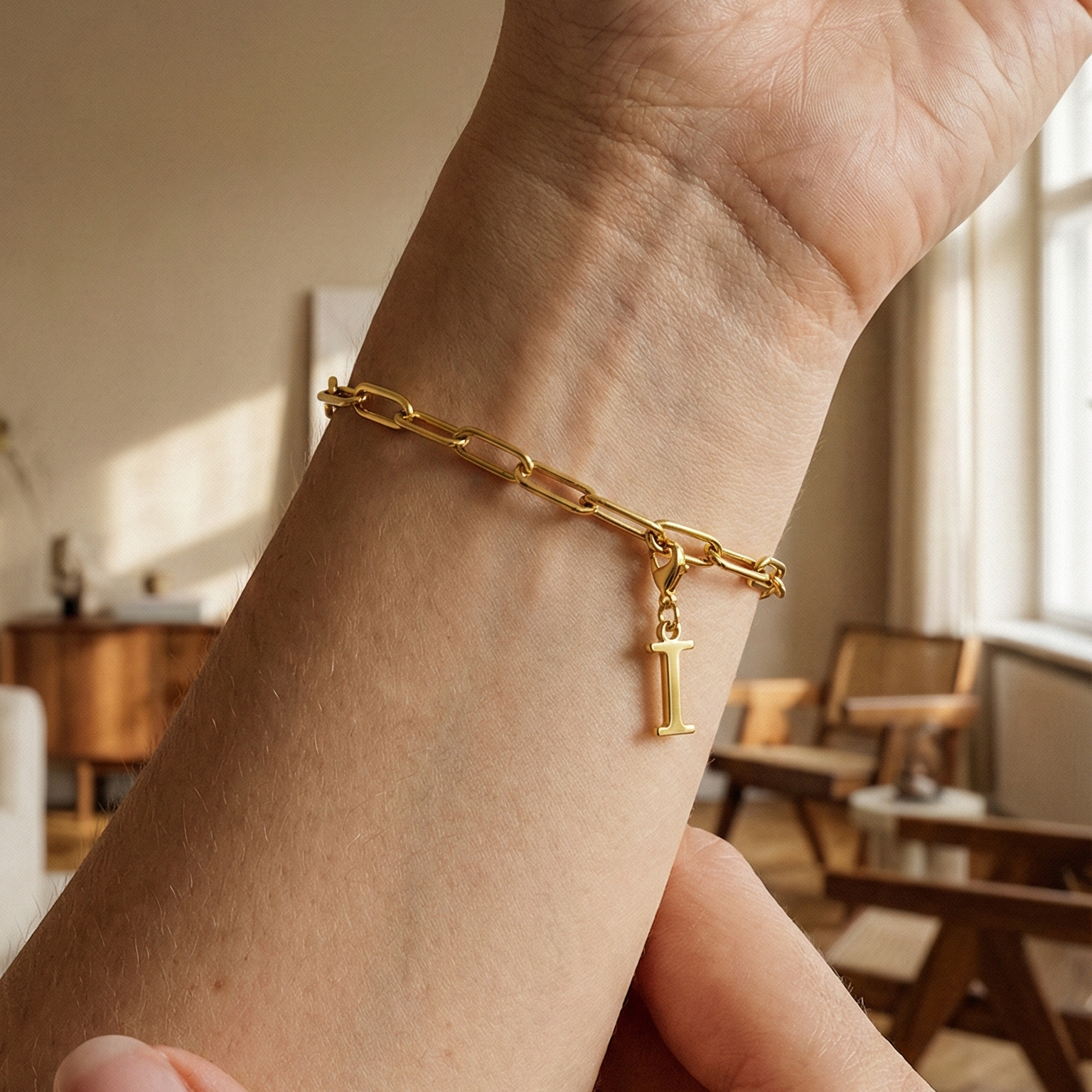 Charm I in gold hängt an goldenem Armband am Handgelenk. Im Hintergrund stylische Altbauwohnung