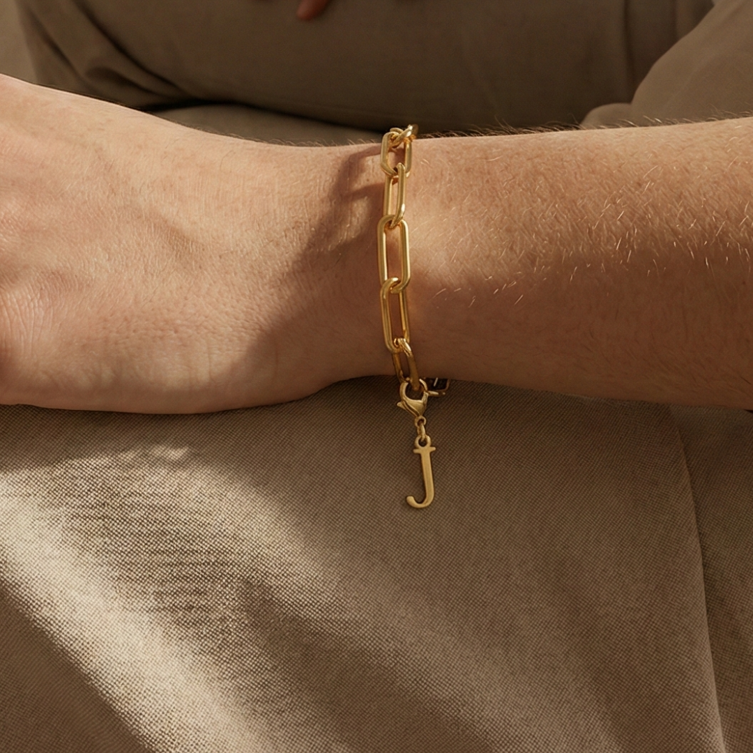 Charm J in gold hängt an goldenem Armband am Handgelenk. Im Hintergrund stylische Altbauwohnung