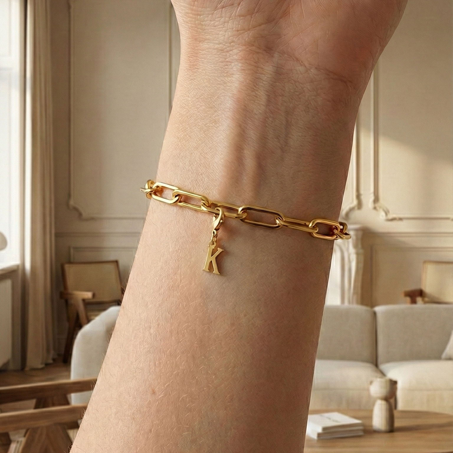 Charm K in gold hängt an goldenem Armband am Handgelenk. Im Hintergrund stylische Altbauwohnung