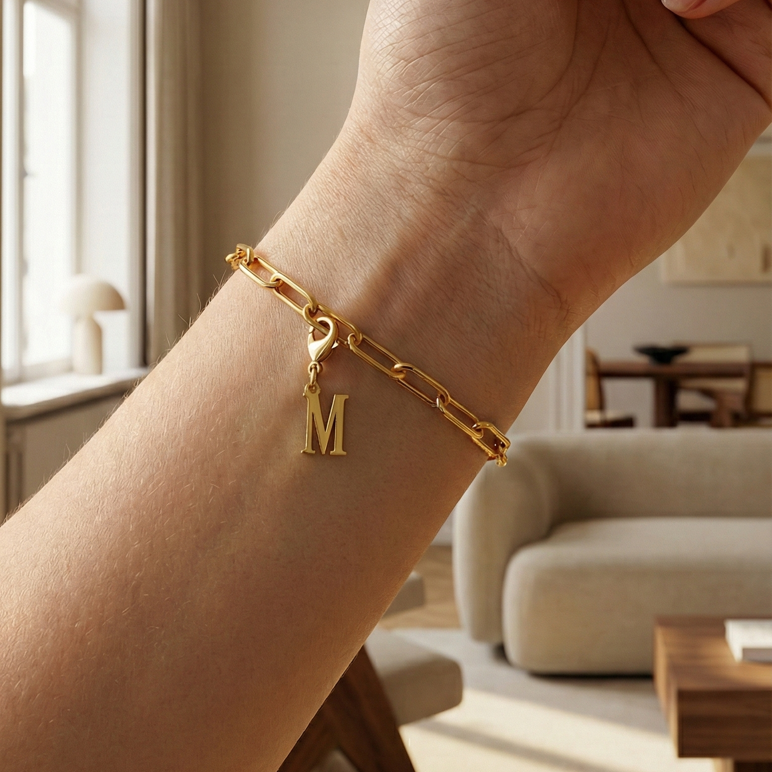 Charm M in gold hängt an goldenem Armband am Handgelenk. Im Hintergrund stylische Altbauwohnung