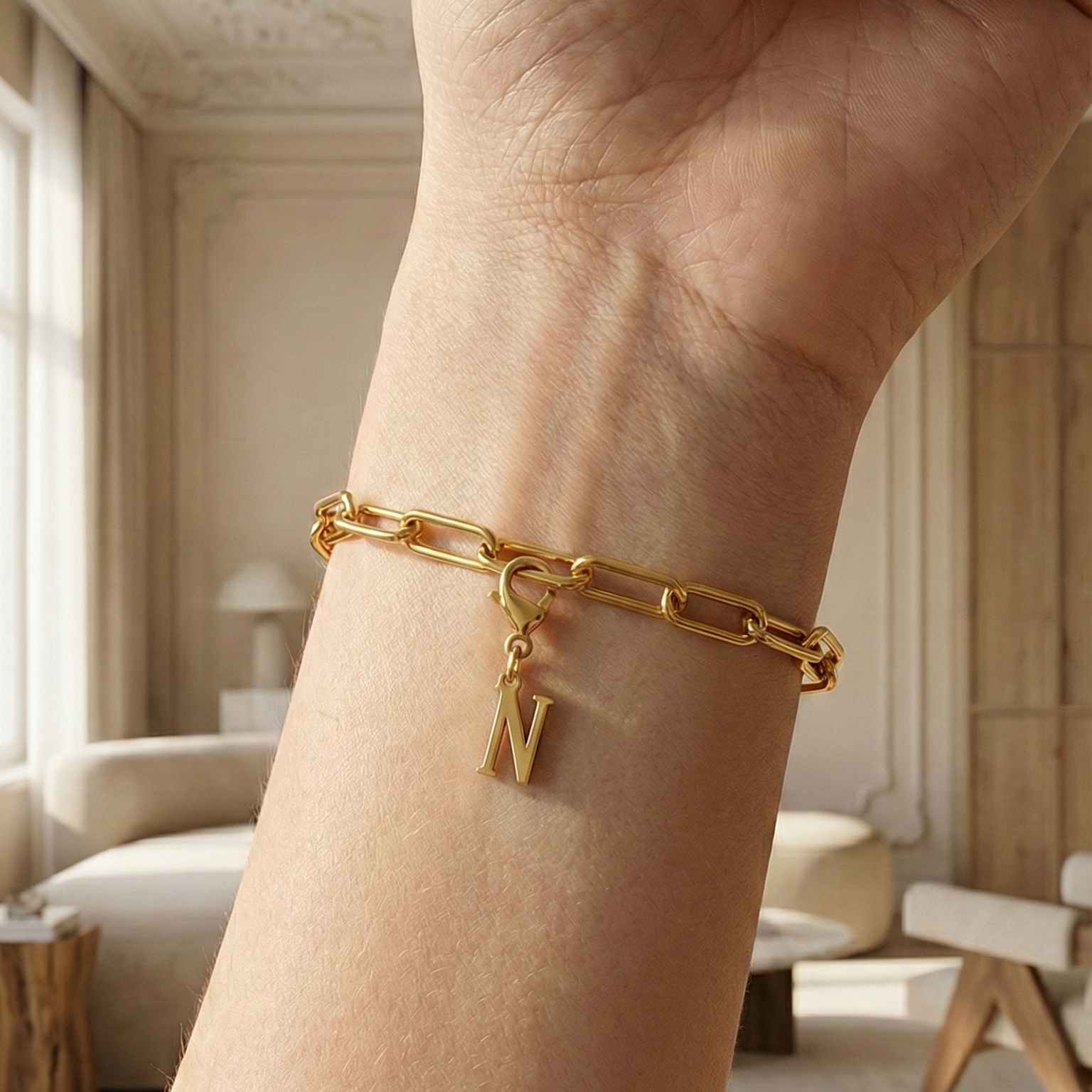 Charm N in gold hängt an goldenem Armband am Handgelenk. Im Hintergrund stylische Altbauwohnung