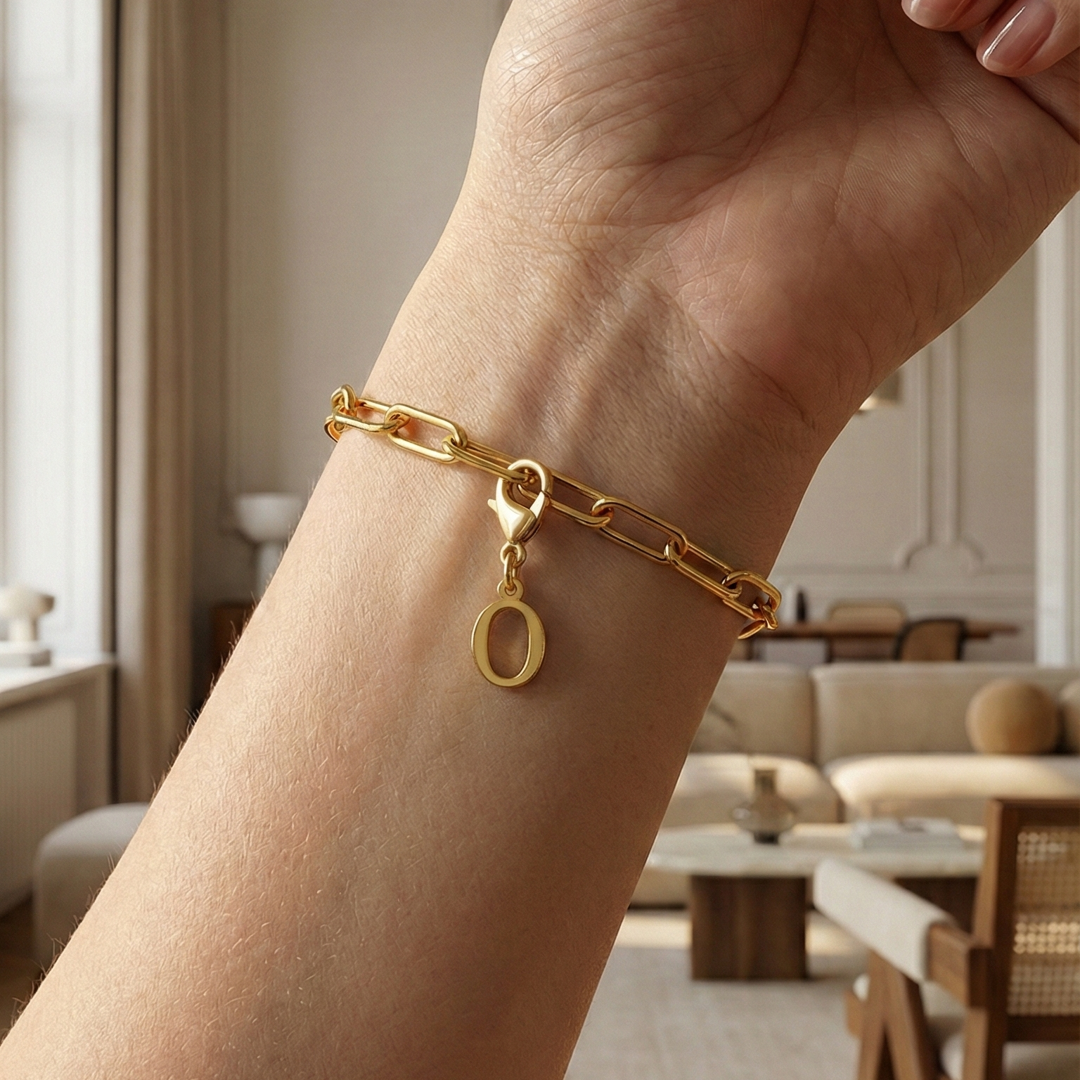 Charm O in gold hängt an goldenem Armband am Handgelenk. Im Hintergrund stylische Altbauwohnung