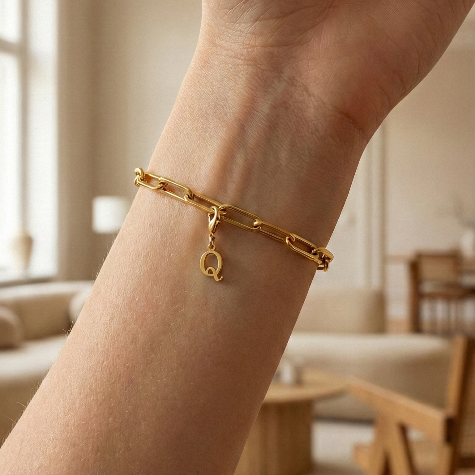 Charm Q in gold hängt an goldenem Armband am Handgelenk. Im Hintergrund stylische Altbauwohnung