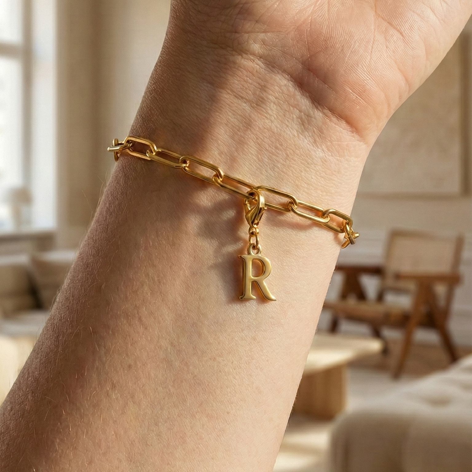 Charm R in gold hängt an goldenem Armband am Handgelenk. Im Hintergrund stylische Altbauwohnung