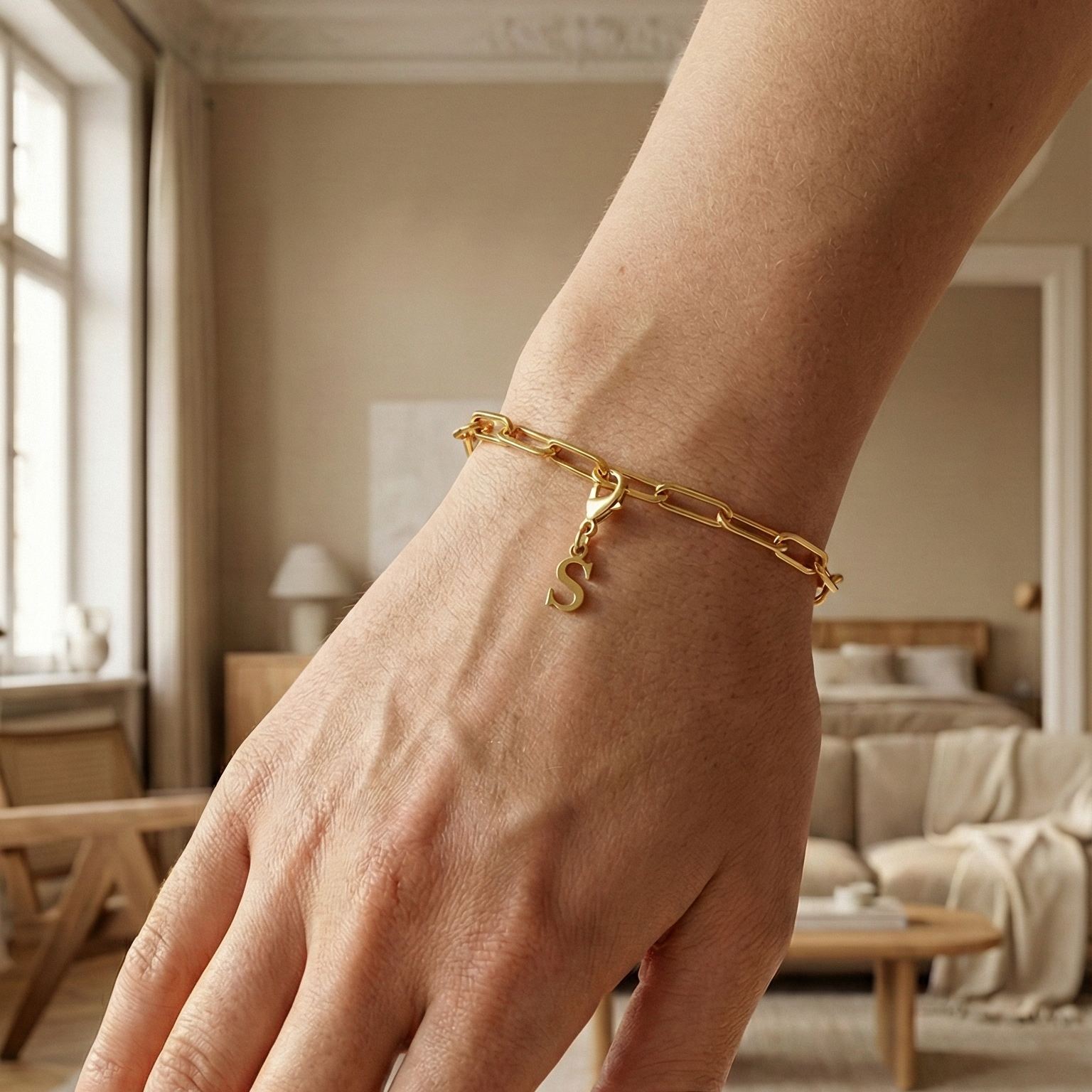 Charm S in gold hängt an goldenem Armband am Handgelenk. Im Hintergrund stylische Altbauwohnung