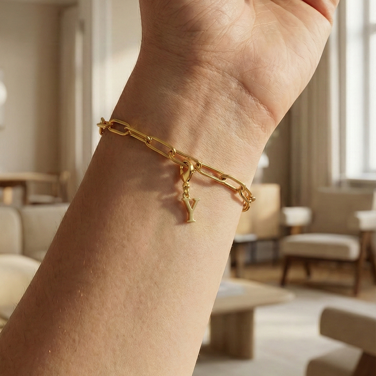 Charm Y in gold hängt an goldenem Armband am Handgelenk. Im Hintergrund stylische Altbauwohnung