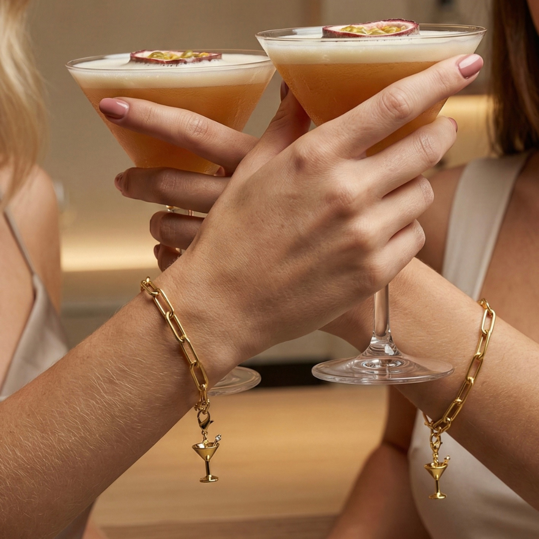 Charm Cocktail in gold hängt an goldenem Armband am Handgelenk der beiden Frauen, die mit Martinis in einer Bar anstoßen. 