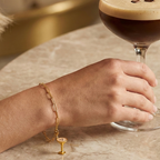 Charm Espresso Martini in gold hängt an goldenem Armband am Handgelenk. der Charm steht auf dem Tisch. Die frau hält das Espresso Martini Glas auf dem Tisch fest