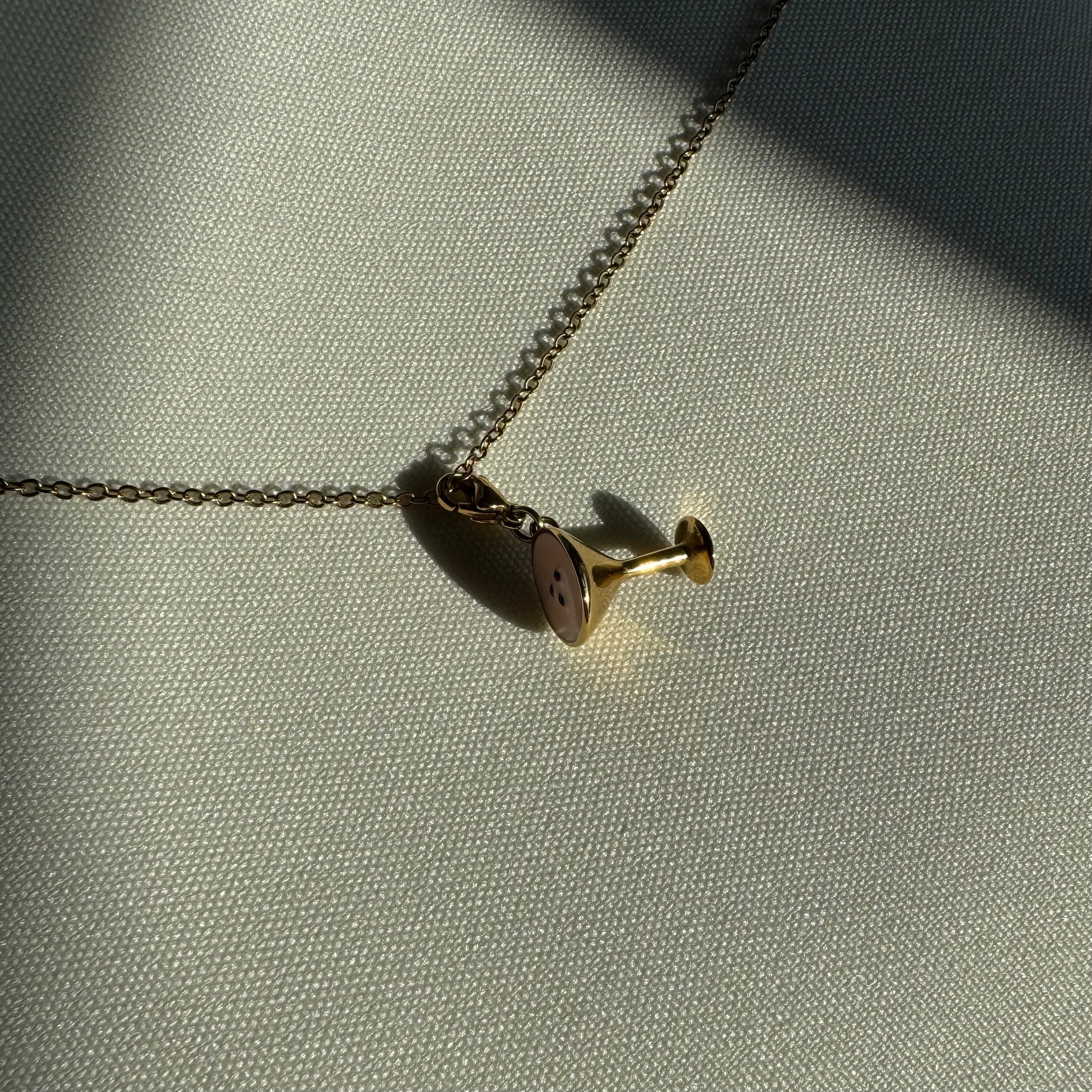 Ein goldener Espresso Martini Anhänger an einer goldenen Kette liegt auf einem weißen Leinentuch. 