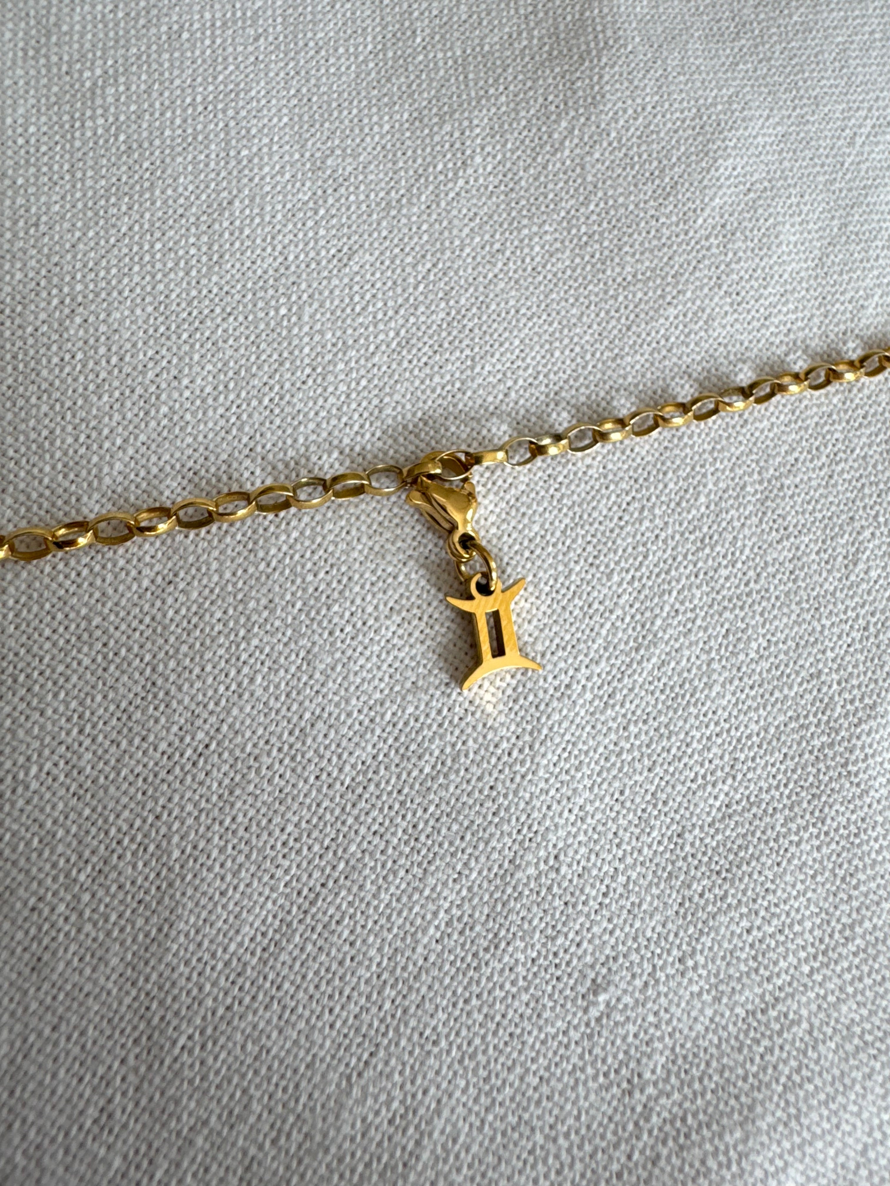 Goldener Zwilling Sternzeichen Charm an  goldenem Armband liegt auf weißem Leinentuch